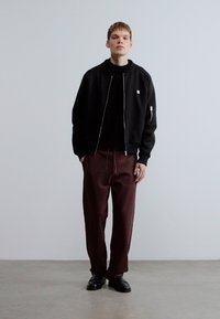 Jachetă bomber neagră cu fermoar peste un pulover negru cu guler înalț, asortată cu pantaloni largi maro și pantofi negri. Design simplu cu detalii funcționale.