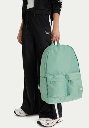 Persona que lleva pantalones deportivos negros de Reebok y zapatillas blancas sostiene una mochila verde claro de Reebok por el asa superior.