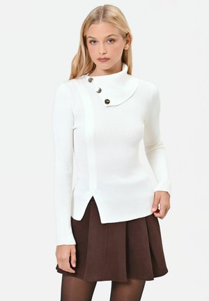 Donna con capelli biondi lunghi che indossa un maglione bianco con collo alto e bottoni e una gonna plissettata marrone scuro su sfondo bianco.