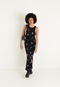 Rochie maxi neagră fără mâneci, cu broderie florală, fante laterale și material texturat. Pur's cu bocanci negri până la gleznă și o poșetă mică.