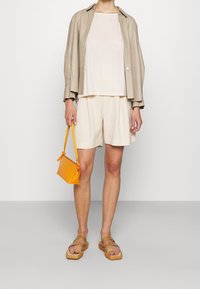 Camicia button-up beige chiaro sopra un top crema e pantaloni corti, abbinata a una piccola borsa arancione e sandali marroni. Design semplice e rilassato.
