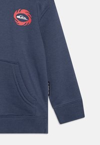 Navy blue sweatshirt with a front pocket, ribbed cuffs, and a circular red logo on the chest featuring a stylized graphic design.