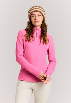 Frau mit pinkfarbenem Fleecepullover und beige gestreifter Strickmütze, steht mit vor dem Körper verschränkten Händen vor neutralem Hintergrund.