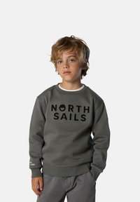 Felpa grigia con logo "NORTH SAILS" in nero. Presenta un colletto a girocollo, polsini e orlo a costine. Indossata sopra una maglietta bianca.