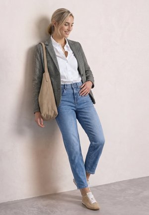 Lachende vrouw leunt tegen een muur, draagt een grijze blazer, witte blouse, blauwe spijkerbroek, beige sneakers en draagt een grote beige schoudertas.