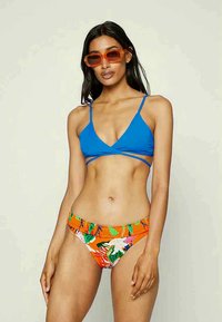 Top de bikini triangular azul com alças finas; parte de baixo de bikini com padrão de flores laranja e cintura franzida, modelo a usar óculos de sol.