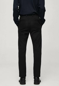 Persoon in zwarte slim-fit broek, zwarte schoenen en een donker lang mouwtop, staand met de rug naar ons toe en met de handen in de zakken.