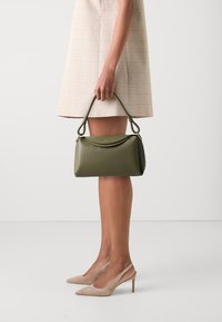 Borsa a tracolla in pelle verde oliva con superficie texturizzata, parte superiore curva e manico corto. Esibita con un vestito a quadri chiari e tacchi beige.