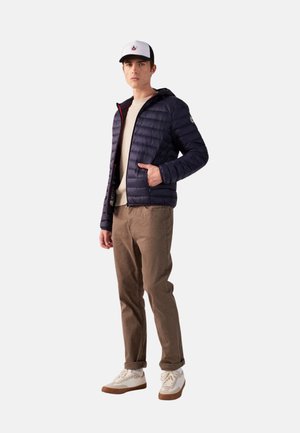 Giovane uomo con giacca imbottita blu navy, camicia beige, pantaloni marroni, scarpe da ginnastica bianche e cappellino bianco e nero, in piedi con una mano in tasca.