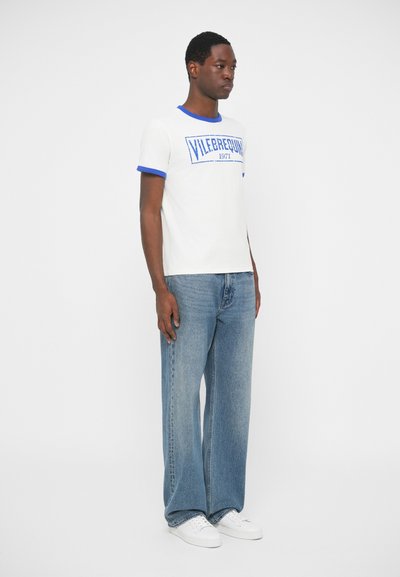 Moški v belem majici z modrim robom in napisom, ohlapnih svetlo modrih kavbojkah ter belih športnih copatih, stoji pred belim ozadjem.