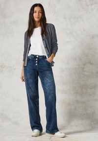 Mörkblå jeans i hög midja med fyra stora knappar, tillsammans med en lätt, texturerad marinblå cardigan och en vit t-shirt.