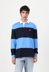 Langermet polo-skjorte i lys og mørk blå striper med hvit krage. Har et lite logo på brystet. Laget av bomull.