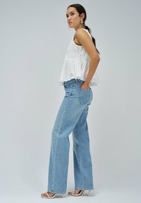 Top blanco sin mangas con pliegues y un dobladillo acampanado, combinado con jeans de pierna ancha azul claro y tacones de dedo abierto blancos, sobre un fondo neutro.