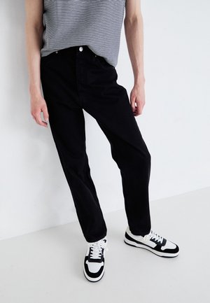 Osoba ubrana w czarne jeansy z wysokim stanem, czarno-białą koszulkę w paski oraz czarno-białe trampki stoi na tle białego tła.