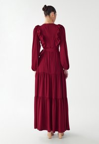 Bordová maxi šaty s dlhými rukávmi, volánkovým výstrihom a vrstveným lemom. Vyrobené z mäkkého materiálu s priliehavým pásom a detailom mašle na chrbte.