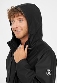 Giacca nera impermeabile con cappuccio, chiusura frontale con zip e cordoncino. Presenta un patch sulla manica con dettagli in bianco.