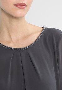 Graue Bluse mit glatter Textur, die einen gefältelten Vorderteil und ein mit Perlen verziertes Dekolleté in Silber und Schwarz aufweist.