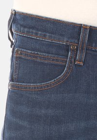 Dżinsy w ciemnoniebieskim kolorze z pomarańczowymi przeszyciami. Posiadają kieszenie, zapięcie na guzik oraz fason slim fit. Widoczna gładka faktura.