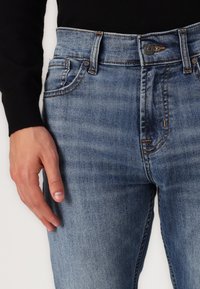 Jeans din denim albastru au un design ajustat, ușor deteriorat, cusături contrastante și un stil cu cinci buzunare, având un sistem de închidere cu un buton.