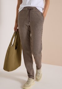 Person wearing brown and white houndstooth pants, beige sneakers, white shirt, holding an olive green large fabric tote bag.