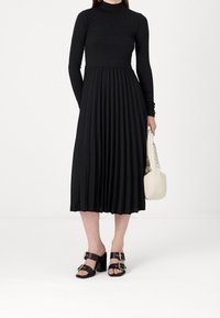Mujer vistiendo un vestido negro de manga larga tipo cuello alto con falda plisada, sandalias de tacón negro con hebillas, sosteniendo un pequeño bolso blanco.