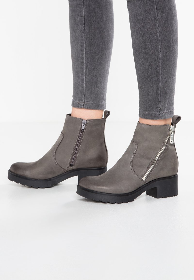 Bottes en cuir gris montant à la cheville avec fermetures éclair latérales et talons épais noirs, portées avec un jean gris foncé ajusté.