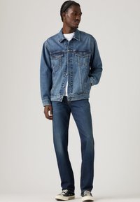 Chaqueta de mezclilla azul con dos bolsillos en el pecho, combinada con jeans azul oscuro. El modelo lleva zapatillas negras y una camiseta blanca debajo.