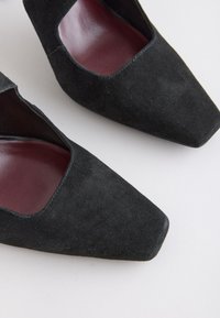 Para czarnych zamszowych balerinek w kształcie kwadratu z bordowymi skórzanymi wkładkami na białym tle.