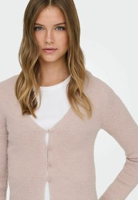 Blush pink knitted cardigan with a V-neck, button closure, and textured fabric. Worn over a white top, featuring long sleeves and soft finish.
