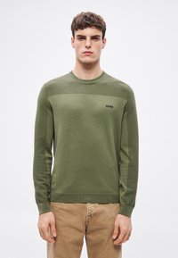 Zöld kötött pulóver kerek nyakkal, a felső részen vízszintes bordázott textúrával, a mellkason pedig egy kis fekete logóval.