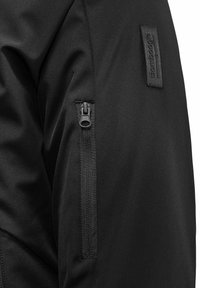 Giacca nera con una texture liscia, dotata di una tasca con zip sulla parte superiore del braccio e di un'etichetta cucita sulla manica.