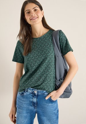 Junge Frau trägt ein grünes, gemustertes Hemd und blaue Jeans, hat einen grauen Rucksack über einer Schulter und lächelt vor einem schlichten Hintergrund.