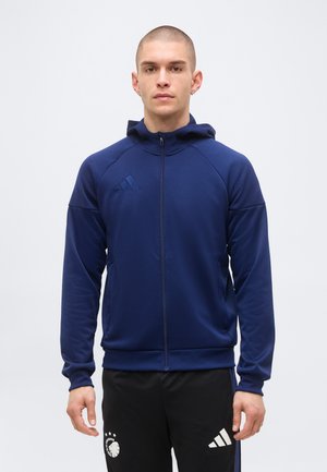 Model bărbat purtând un hanorac bleumarin cu fermoar și pantaloni sport negri Adidas cu logo-uri sportive, în picioare pe un fundal simplu și deschis.