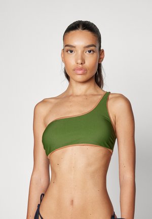 Frau mit langen dunklen Haaren, die einen olivgrünen Bikini mit einer Träger auf der Schulter und hellbraunen Besatz trägt, vor einem schlichten hellen Hintergrund.