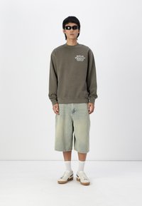 Sudadera verde oliva con texto frontal, pantalones cortos de mezclilla azul claro, y calcetas blancas con zapatillas que tienen una suela marrón y negra.