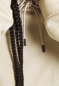 Beige mikina s texturovanou tkaninou, vybavená pletenými šňůrkami s černými špičkami probíhajícími výstřihem.