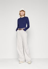 Top de manga larga azul marino, pantalones de pierna ancha beige, cinturón marrón y gorra blanca. Tela suave con un corte relajado; los zapatos tienen un suave tono crema.