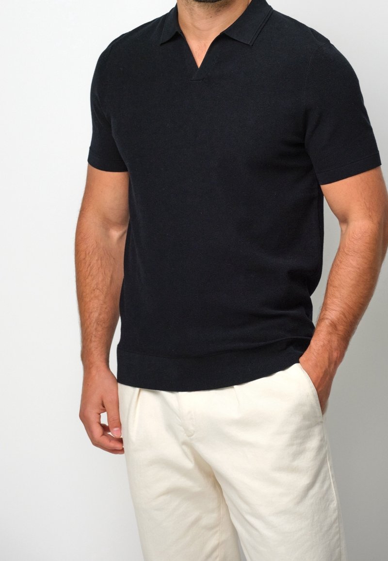 Homme portant un polo noir à manches courtes et un pantalon écru, debout avec une main dans la poche devant un fond uni.