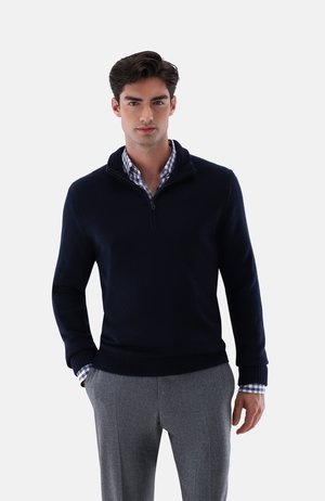 Marinefarbener Zip-Pullover mit gerippten Bündchen und Saum, weicher Textur. Getragen über einem hellblauen und weißen karierten Hemd mit hochgekrempelten Ärmeln.