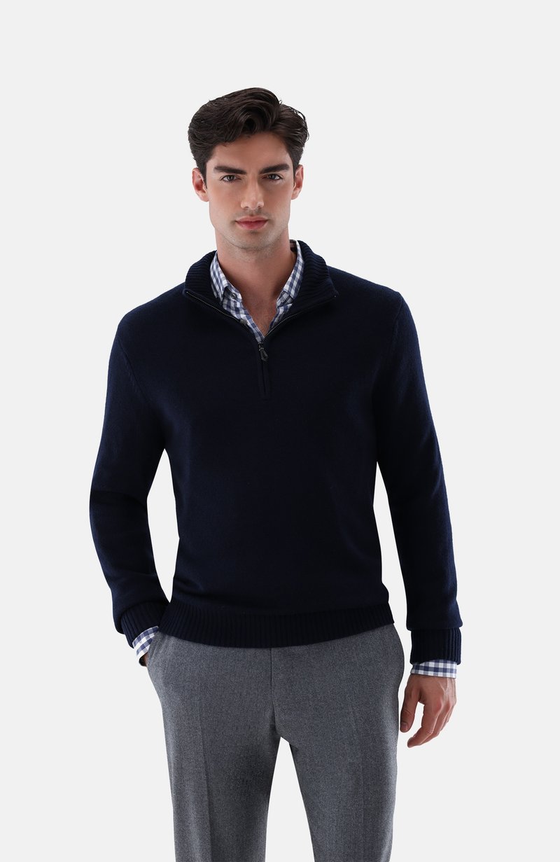 Marinefarbener Zip-Pullover mit gerippten Bündchen und Saum, weicher Textur. Getragen über einem hellblauen und weißen karierten Hemd mit hochgekrempelten Ärmeln.