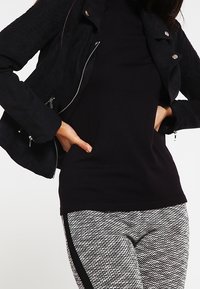 Veste noire texturée avec des zips et des boutons-pression, portée avec un haut noir ajusté et un pantalon à motifs gris et blanc avec des rayures noires sur les côtés.