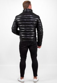 Giacca in pelle nera imbottita con colletto alto, texture a costine e design attillato, abbinata a jeans neri e scarpe bianche.
