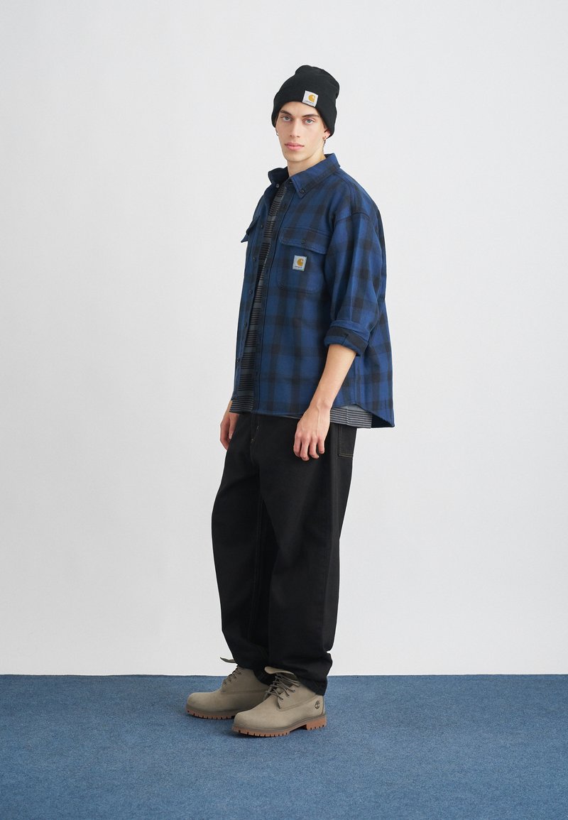 Blauwe en zwarte geruite flanellen shirt, grijs gestreept T-shirt eronder, zwarte wijde broek, en grijze laarzen met bruine zolen, zwarte beanie.