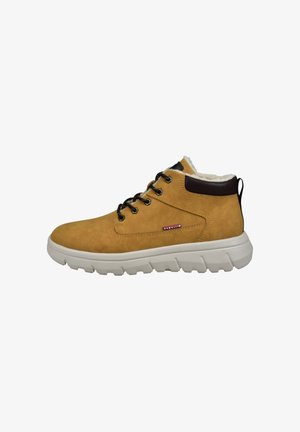 Helle Wildleder-Mid-Top-Schuhe mit einer weißen Gummisohle, schwarzen Metall-Schnürschlaufen und einem mit Fell gefütterten Kragen mit einem braunen Akzent oben.