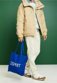 Beige gepolsterte Jacke mit grünen Kordeln, hellblauer Strickpullover, cremefarbene Hose und blautasche mit weißem "ESPRIT"-Logo. Legere Sportschuhe.