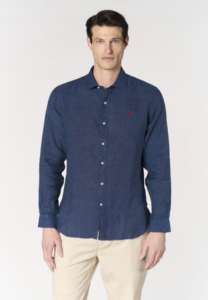 Uomo che indossa una camicia slim fit blu scuro con bottoni e maniche lunghe e pantaloni beige, in piedi su uno sfondo bianco uniforme.