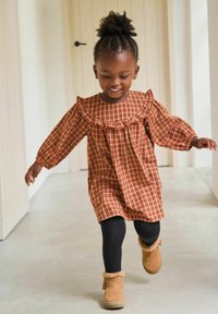 Kind in einem rot-beigen karierten Kleid mit Rüschensaum und langen Ärmeln, schwarzen Leggings und braunen, fleecegefütterten Stiefeln, das drinnen geht.