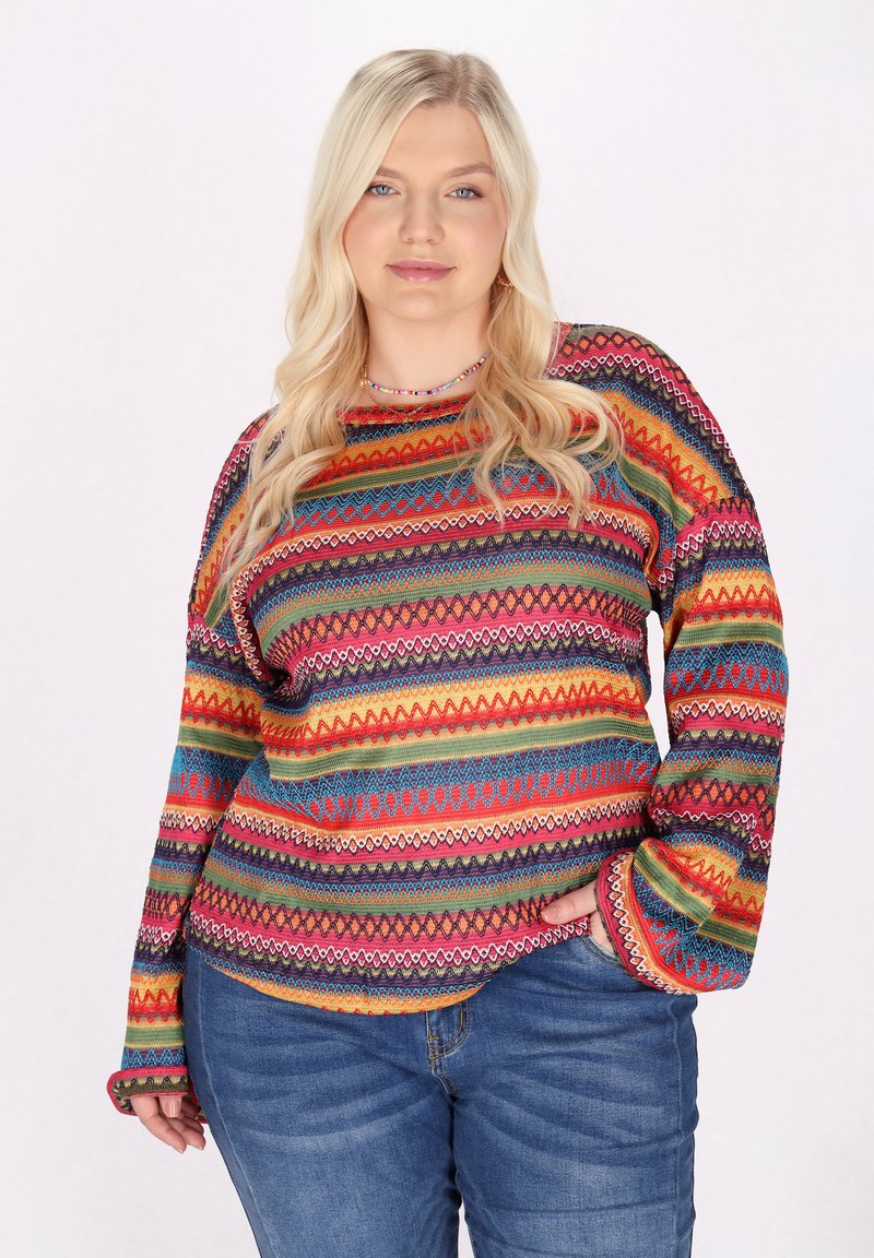 Buntgestreifter Pullover mit lebendigen Zickzack- und geometrischen Mustern, aus einem strukturierten Strickstoff und mit langen Ärmeln. Kombiniert mit Jeans.