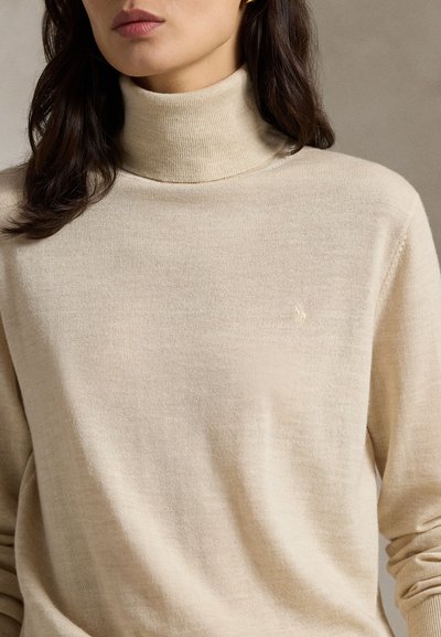 Kvinne iført en beige genser med høy hals, langt mørkt hår, synlige lepper og en subtil logo brodert på brystet.