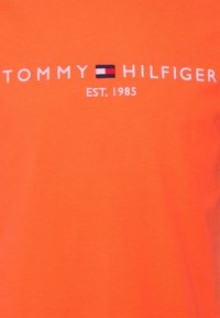 Oranžové bavlněné tričko s bílým vyšívaným textem "TOMMY HILFIGER EST. 1985" a barevným blokovým logem ve středu.